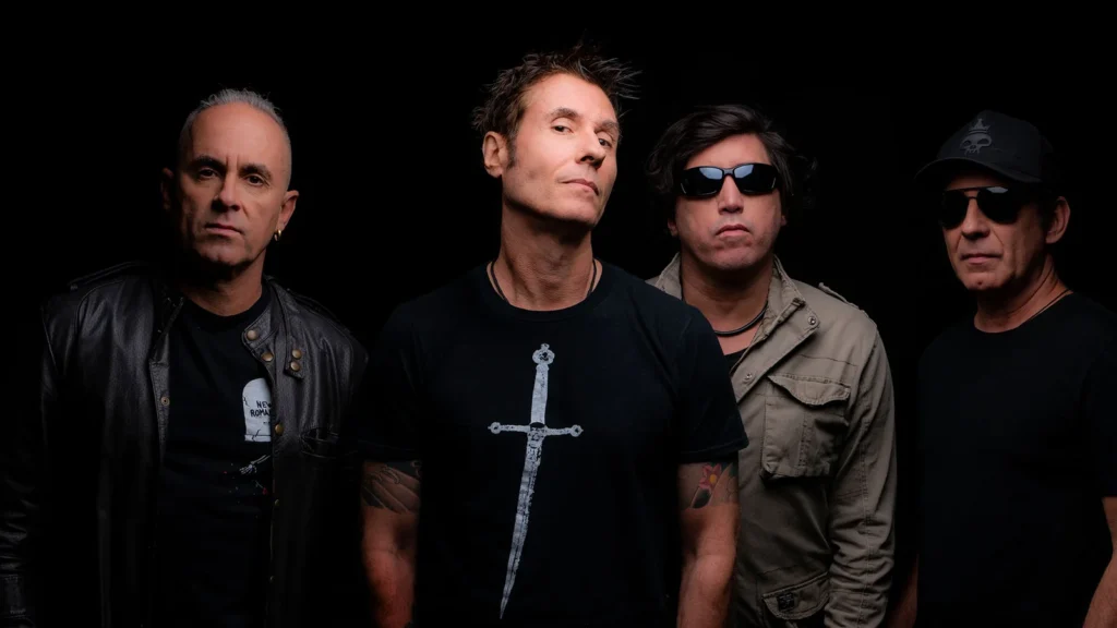 Fotografia promocional da banda de rock brasileira Capital Inicial. Os quatro integrantes estão posicionados lado a lado contra um fundo preto sólido. À esquerda, um integrante usa uma jaqueta de couro preta sobre uma camiseta escura. Ao centro, o vocalista Dinho Ouro Preto veste uma camiseta preta com a estampa de uma espada vertical. À sua direita, o terceiro integrante usa uma jaqueta estilo militar na cor cáqui. Na extremidade direita, o quarto integrante usa óculos escuros, camiseta preta e um boné escuro com o logotipo de uma caveira coroada. Todos mantêm expressões sérias e uma postura clássica de banda de rock.