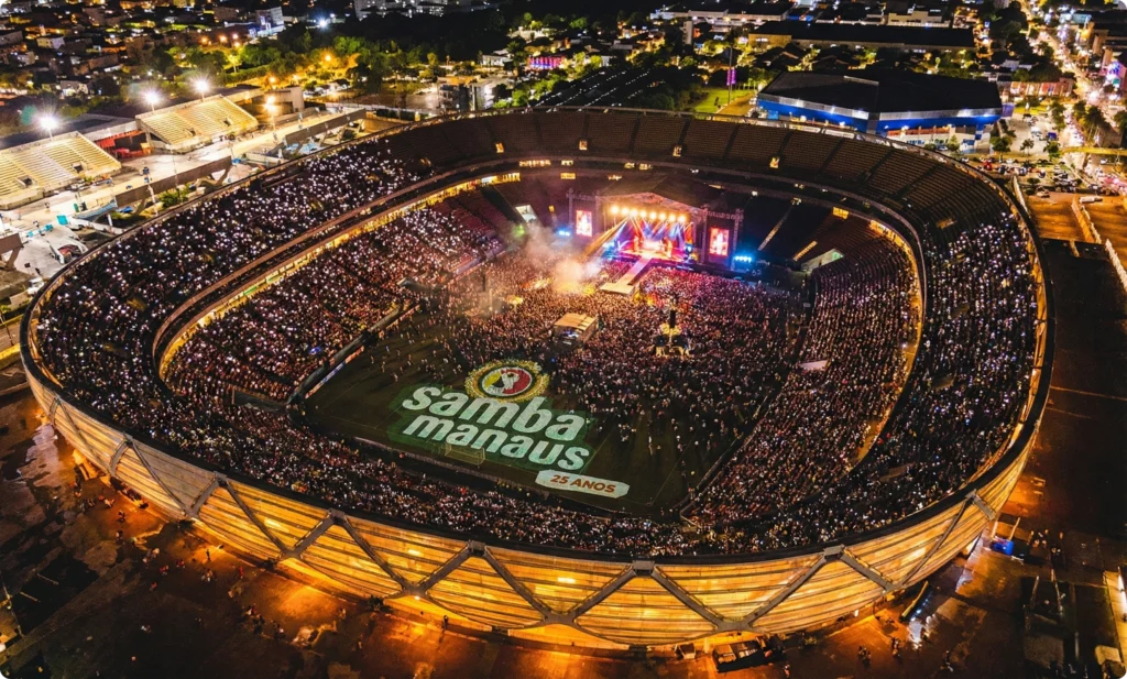 Fotografia aérea noturna em ângulo picado da Arena da Amazônia, em Manaus, totalmente iluminada e lotada para o evento Samba Manaus. O estádio, com sua arquitetura icônica inspirada em um cesto indígena, brilha em tons dourados sob as luzes da cidade ao fundo. No gramado, uma multidão imensa se concentra diante de um palco monumental com luzes vibrantes, telões de LED e efeitos de fumaça. No centro do gramado, destaca-se uma projeção ou aplicação gráfica em grande escala do logotipo 'Samba Manaus 25 Anos', com o ícone do tucano e as cores verde e laranja. As arquibancadas estão repletas de milhares de pequenos pontos de luz vindos dos celulares do público, criando um efeito de mar de estrelas dentro da arena.