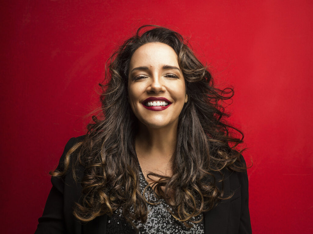 Retrato de rosto da cantora Ana Carolina sorrindo alegremente. Ela possui cabelos castanhos longos e ondulados que emolduram seu rosto. A artista usa batom vermelho escuro e uma blusa preta com brilhos. O fundo é de uma cor vermelha sólida e vibrante, criando um contraste marcante com sua imagem